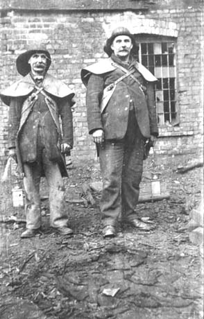 Photograph showing two men dressed in long jackets, sou'westers, and with leather capes strapped over their shoulders and backs; each is holding a miner's lamp; behind the men, is a brick building with a window; discarded material is lying round their feet; they have been identified as Bert Henderson, Senior, and Bill McKeand, shaftsmen at Wheatley Hill Colliery