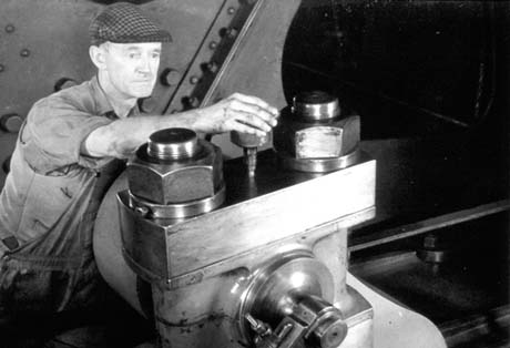 Photograph showing a man wearing a shirt, waistcoat and cap, behind a large piece of machinery which appears to have outsize nuts and bolts on it; he is screwing a round cap with a pointed stem into place; the cap has been identified as a grease Cup and the man as Bert Henderson of Wheatley Hill