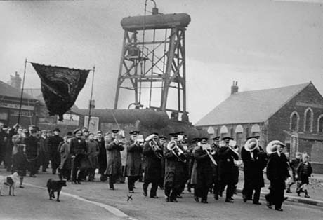 Photograph showing the members of a band walking along a road playing; they are being followed by men carrying a lodge banner; behind the procession a large cage-like structure can be seen; beyond that the side and end of, most likely, a non-conformist church can be seen; the photograph has been identified as Band at Vesting Day, Wheatley Hill; Vesting Day was 1st January 1947, when the coal industry was nationalised