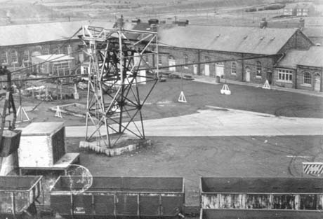 Photograph showing, from above, two single-storey buildings forming a right-angle, with buildings and excavations in front of them; also in front of them is an overhead pulley system supported by a pylon-like structure; in the foreground are empty coal trucks; in the distance, beyond the buildings, are open fields; the photograph has been identified as No. 2 Winder To Work joiners and Fitters, Wheatley Hill