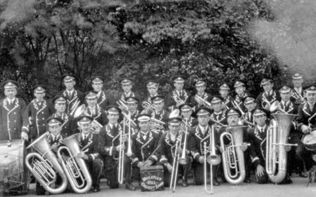 Photograph showing twenty five men and three boys in uniform and holding musical instruments, posed against trees; in front of a man on the front row is a box carrying the words: Wheatley Hill Colliery Band; the man behind the box may be the leader of the band who has been identified as Bob Walker