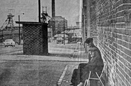 Photograph showing, on the right, a wall with two elderly men sitting against it; in front of the men are a wide pavement, a brick bus stop and a road running away from the camera; at the end of the road the winding gear, winding house, chimney and low buildings of a colliery can be seen; a car is moving on the road away from the camera and a bus is approaching the camera; the photograph has been identified as Front Street with Colliery in Background, Wheatley Hill