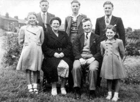Photograph showing a woman wearing an overcoat sitting with a man wearing a dark jacket, tie, jumper and light trousers; behind them three young men wearing jackets and ties are standing; on the left a girl, aged approximately ten years and wearing an overcoat is standing; on the right, a girl, aged approximately seven years and wearing an overcoat, is standing; they are posed in a garden with houses in the background; they have been identified as Ralph and Connie Woodland with Family in Wheatley Hill