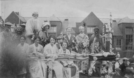 Photograph showing seven women and six men in costume, possibly on a carnival float, with what appears to be a school building behind them; four of the women are wearing kimono-type garments and one is wearing a bonnet; two men are dressed as Chinamen and two are wearing Cossack-type garments; two other men are wearing kimono-type costume; at the right of the picture part of a crowd can be seen watching something out of range of the camera to the right; the people in the centre of the photograph have been identified as Operatic Society, Wheatley Hill
