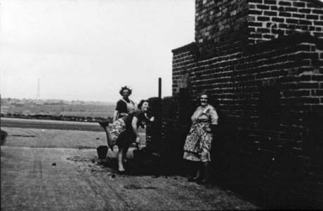 Photograph showing three women in dresses and pinafores by the side of a house with countryside behind them; one woman is standing leaning against the wall; another is leaning forward and kicking up her right leg; the third is standing behind the second woman looking towards the camera; the photograph has been described as Putting in The Coals, Wheatley Hill; it is possible that the women are shovelling coal into a coal hole in the wall, but the photograph is too dark for that to be seen