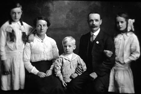 Photograph showing a girl, aged approximately twelve years, wearing a light-coloured dress, standing on the left of the photograph, resting her arm on the shoulder of a woman, who is wearing a light blouse and dark skirt; next to her, in the middle of the photograph, is a boy, aged approximately six years, wearing a sailor suit; his arm is being held by a man wearing a dark suit, tie and stiff collar; on the right is a girl aged approximately nine years with her arm through that of the man; they have been identified as The Henderson Family of Wheatley Hill