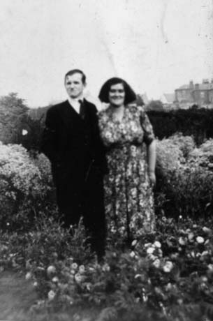 Photograph showing a man and a woman standing facing the camera in a garden with blooming plants in front and behind them; the man is wearing a dark suit and the woman a flowered dress; they have been identified as Steve and Vera Woodley, formerly Howie, of Wheatley Hill