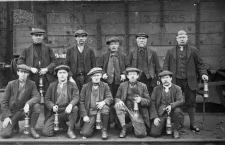 Photograph of five men standing in front of a coal waggon with five men kneeling on the ground in front of them; they are all dressed in caps, jackets, ties or scarves, waistcoats, trousers or leggings and boots; all are carrying miner's lamps; they have been identified as Waggon Waymen, Wheatley Hill