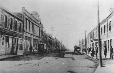 Photograph showing a road running away from the camera; on the left of the picture, a large department store can be seen with indistinct buildings beyond; the name of the department store cannot be discerned; on the right of the picture, the road is lined with the facades of terraced houses; indistinct figures can be seen on the right-hand pavement; a motor van can be seen parked facing away from the camera; a horse-drawn cart can be seen facing away from the camera on the right of the picture; the photograph has been identified as Front Street From Nimmo, Wheatley Hill