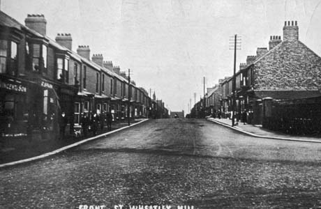 Postcard photograph entitled Front Street, Wheatley Hill, showing a road running away from the camera; a shop with the name, Henderson, can be seen near the camera on the left, but most of the left of the street appears to be terraced houses with bay windows; the right of the street also appears to consist of terraced houses with bay windows; a church can be seen at the end of the street in the distance