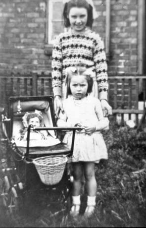Photograph showing a small girl, aged approximately four years, wearing a short light-coloured frock, standing at the handle of a doll's pram with a doll inside it; behind her is another girl, aged approximately twelve years, wearing a patterned jumper; behind them is part of a brick house; they have been identified as Edith and Margaret Dixon of Wheatley Hill
