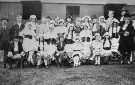 Photograph of twenty six people in costume posed in front of a low wooden building; they are dressed as peasant girls, courtiers, soldiers and clergymen; they have been identified as members of the Wheatley Hill Operatic Society