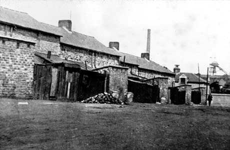 Photograph showing the backs of three terraced houses showing privies and fences; in the distance, at the right of the photograph, the winding gear of the colliery can be seen; the photograph has been described as Slum Clearance, Wheatley Hill