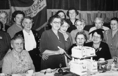 Photograph showing nine middle-aged women standing behind three other middle-aged women sitting at a table on which crockery can be seen; a middle-aged woman is standing at the table holding a knife with which she is cutting a ceremonial iced cake of two tiers; behind the women is a banner reading: British Legion Women's Section Wheatley Hill Branch