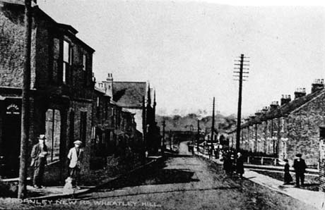 Postcard photograph entitled Thornley New Road, Wheatley Hill, showing a road running away from the camera with terraced houses on the right and what appears to be a railway bridge over the road in the distance; an unidentifiable shop is on the corner of the left side of the road with, possibly other shops further down the road; unidentifiable figures can be seen on the pavements