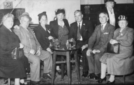 Photograph showing three middle-aged men and four middle-aged women sitting in a semi-circle round a small table on which three glasses can be seen; the men are wearing suits and the women overcoats and hats; the photograph has been identified as having been taken in Bobby Winters Pub Over The Beck, Wheatley Hill