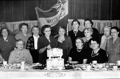 Photograph showing nine middle-aged women standing behind three other women sitting at a table on which crockery can be seen; a woman is standing at the table holding a knife with which she is about to cut a ceremonial iced cake of two tiers; behind the women is a banner reading: British Legion Women's Section Wheatley Hill Branch