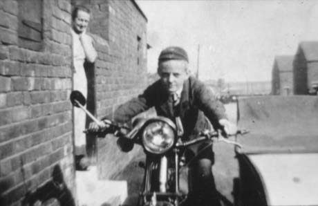 Photograph of a boy aged approximately twelve years, wearing a school cap, sitting astride a motor bicycle and looking at the camera; he is being watched by a woman in a pinafore who is standing in the opening in a wall leading to a yard; beyond the boy, the ends of terraces can be seen; the photograph has been identified as Mr. W. Henderson and Mother in Ford Street, Wheatley Hill