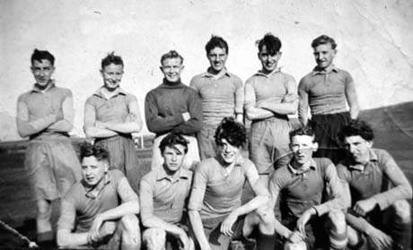 Photograph of eleven young men in football strip posed on a football field in what appears to be a high wind; they have been identified as being in Wheatley Hill