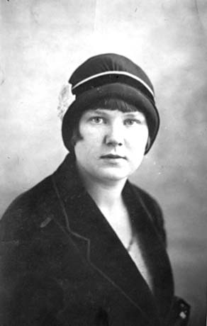 Photograph of the head and shoulders of a young woman wearing a dark hat with a light ornament in it on the left; she has short dark hair showing below her hat; she is wearing a dark coat or jacket over a dress; she has been identified as Meg Rutherford of Wheatley Hill