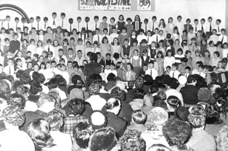 Photograph showing a choir of approximately one hundred and thirty children facing the camera with an audience of approximately seventy adults looking at the choir with their backs to the camera; on the wall behind the choir is a notice reading: School Music Festival 1969; the photograph has been described as Wheatley Hill and Shotton Music Festival