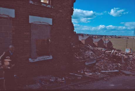 Photograph showing the exterior of a terraced house with its windows and door bricked up; the house next to it has been demolished and is a heap of bricks, beyond which the ends of three terraces can be seen; a man is standing in front of the bricked-up house carrying a pile of wood; the photograph has been identified as Demolition of Numbered Streets, Wheatley Hill
