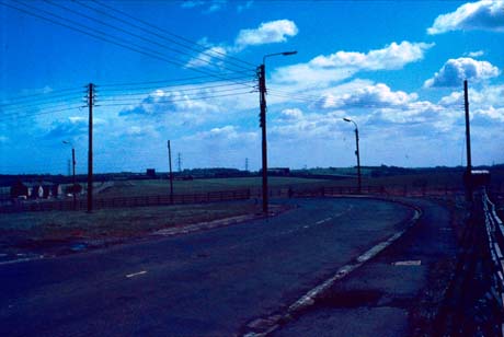 Photograph showing a road curving to the left with an open field beyond it; small huts can be seen on the left of the picture; the photograph has been described as showing the site of the colliery at Wheatley Hill