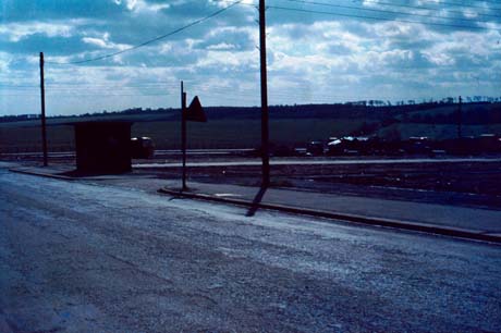 Photograph showing the surface of a road in the foreground with indistinct cars parked at the right of the picture; in the centre in the distance is an indistinctly seen open field which has been identified as the site of the former colliery at Wheatley Hill