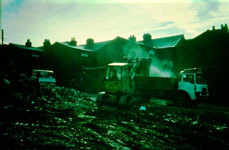 Photograph showing two lorries and a vehicle with caterpillar tracks scooping bricks into one of the lorries; behind the vehicles the backs of terraced houses can be seen; in the foreground are bricks lying on the ground; the photograph has been identified as showing The Demolition of Numbered Streets in Wheatley Hill