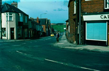Photograph showing a road running away from the camera to the right; at the end of the street, fields can be seen; an unidentifiable shop is on either corner of the street and terraced houses can be seen on the left-hand side of the picture; a man is walking up the street and the rear of a car can be seen; the street has been identified as Thornley Road, Wheatley Hill