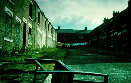 Photograph showing two rows of terraced houses with another row across the end; the houses on the left of the photograph can be seen to be bricked up, although a line of washing can be seen further along the terraces; the area in front of the boarded-up terraces is overgrown and a chair lying on its side is at the front of the photograph; the photograph has been identified as Demolition of Numbered Streets, Wheatley Hill