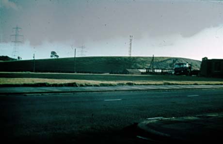 Photograph showing the surface of a road at the front of the picture; across the road an open field with low hill behind it can be seen; the photograph has been identified as Reclamation of Pit Head, Wheatley Hill; at the right of the picture a lorry and what appears to be a line ot coal trucks can be seen near the low hill
