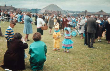 Photograph showing the backs of two women kneeling on the ground taking a photograph of a small boy dressed as a pierrot and of a small girl dressed as a shepherdess; the women and children are part of a crowd of people on a field with semi-detached houses on its edge; behind the houses what appears to be a pit-heap can be seen and, behind that, further houses in the far distance; the photograph has been described as Carnival, Wheatley Hill