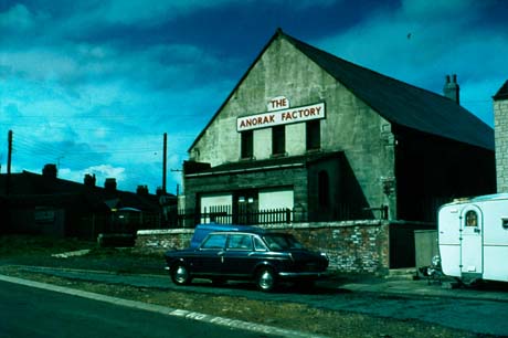 Photograph of the exterior of a building with a sign reading The Anorak Factory on its end; below the sign are three narrow windows and, below them, a wide porch; a low wall is in front of the building; in front of the wall a van, a car and a caravan are parked; the roofs of houses can be seen on the left of the picture; the photograph has been identified as Chapel, Now Anorak Factory, Wheatley Hill