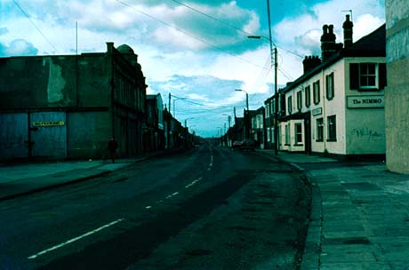 Photograph showing a road running away from the camera; on the right a public house called The Nimmo can be seen with indistinct buildings further along the street; on the left of the picture a large indistinct building with a No Parking sign on it can be seen; the buildings beyond can be seen only indistinctly; the photograph has been identified as Front Street, Wheatley Hill