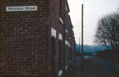 Photograph showing part of the end of a terrace of houses with the name Weardale Street on it; the front of the houses can be seen obliquely, as can the road in front of them; a car is parked half way down the road; beyond the road is open country; one house appears to be bricked up; the photograph has been identified as Weardale Street- Last To Remain, Wheatley Hill