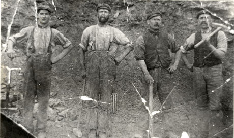 Photograph of four men wearing work clothes of trousers, shirts, waistcoats and caps standing in front of a rock face; the man on the left is holding a fork, the man next to him is holding a hammer as are the other two men in the picture; they have been identified as stone quarry workers in Wingate