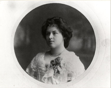 Photograph of the head and shoulders of a young woman with dark hair dressed in a roll round her head; she is wearing a necklace close round her throat and a lace dress with a flower pinned on her left shoulder; she has been identified as Eva Russell of Wingate