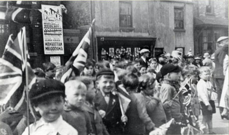Photograph of a crowd of children, aged approximately between six and ten years, carrying Union Jacks; approximately seven adults can be seen at the right of the picture; the children are in front of an unidentifiable shop, described as Morrisons, with bunting in its window; behind the children, also, is a poster advertising the programme of The Palace Cinema, Wingate; the films advertised are Cockeyed Cavaliers and Clark Gable in Manhattan Melodrama; it is possible that the occasion being marked is the Silver Jubilee of George V in 1935