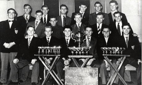 Photograph of eighteen young men dressed in suits and ties posed against a wall; two of them are wearing blazers bearing a badge; in front of them are three card tables on which there are small trophy cups and one large cup; in front of the tables is a black board on which is written the following: Wingate Wanderers Champions Hartlepool and District League Div II 1956-57 Finalists ....