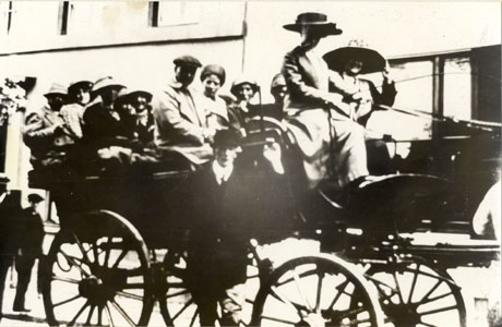 Photograph of a large four-wheeled vehicle drawn by a horse which is out of shot; there are approximately eleven people in the vehicle dressed in overcoats and hats; a man is standing in front of it and two men can be seen standing in its rear; it has been identified as The Brake, Wingate