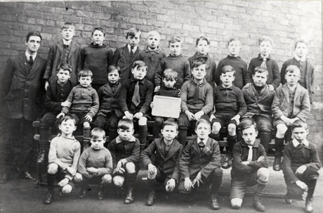 Photograph of twenty five boys aged approximately ten years posed against a wall; a man is standing with them; a child is holding a notice which cannot be read; they have been identified as pupils from a school in Wingate