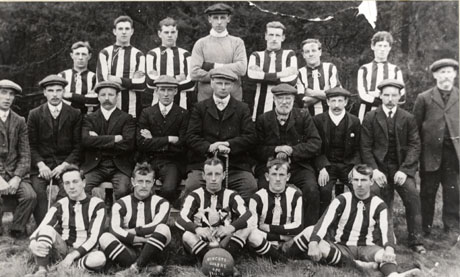 Photograph of twelve men in football strip posed with nine men in suits and caps; a football at the front of the picture bears the words Wingate Quarry A[thletic] F[ootball] C[lub] 1912-13