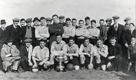 Photograph showing eleven men in football strip in a group with fifteen other men in suits and overcoats; they are also accompanied by a woman with a small boy aged approximately seven years;there is a large trophy cup at the front of the group; they have been identified as the Wingate Welfare Football Club