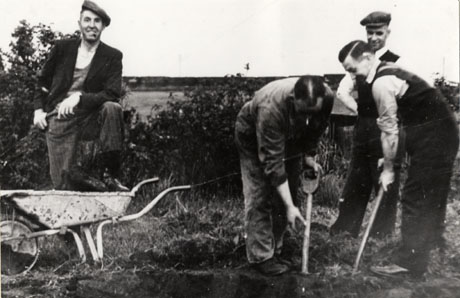 Photograph showing a man at the left of the picture standing in front of bushes with a field beyond; he is wearing a jacket, trousers and a cap and is standing with his foot on a wheelbarrow; on the right of the picture are two men digging with spades and a third man is watching them; one of the men digging is wearing overalls and the other two are wearing trousers and waistcoats; they have been identified as working on the Welfare Park in Wingate