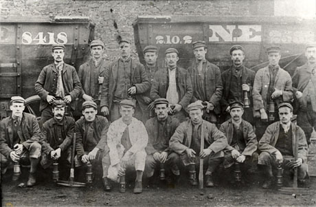 Photograph showing nine men standing, and eight men squatting, in front of two coal waggons, which bear the numbers 8418 and 2. 10. 2 and 2532; all the men are holding a miner's lamp and three of the men on the front row are holding picks; they are all dressed in work clothes of cap, jacket, waistcoat scarf and boots; they have been identified as miners at Wingate