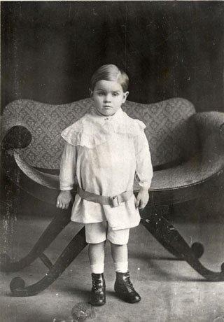 Photograph of a small boy aged approximately five years standing in front of a chair; he is dressed in a tunic with a large lacey collar and a belt round it, a pair of short trousers, button boots and socks; he has been identified as Mr. Walter Bell of Wingate