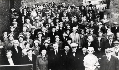 Photograph of approximately one hundred and twenty elderly men and women, standing on grass near the wall of a building, photographed from above; a car can be seen at the rear of the group on the right; they are dressed in overcoats, suits, cardigans and dresses; they have been identified as taking part in an Over 60s Party in Wingate
