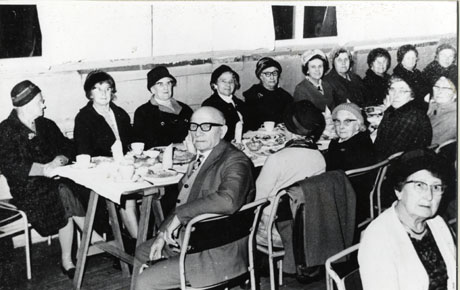 Photograph of ten elderly women sitting with their backs to the wall at a long table; four elderly women and one elderly man are sitting opposite them with their backs to the camera; the man has, however, turned round to face the camera; the face of one woman sitting at another table parallel to the first can also be seen; all the women are wearing hats and most are wearing overcoats; they have been identified as members of the Over 60s Club in Wingate