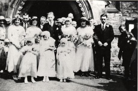 Photograph showing a newly married couple standing outside the door of a church, accompanied by one adult bridesmaid, three child bridesmaids, and thirteen adults most of whom are standing behind them; the photograph has been identified as recording J. Cowie's Wedding